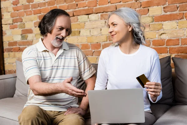 Femme heureuse et mature tenant carte de crédit près de mari d'âge moyen pointant avec la main à l'ordinateur portable — Photo de stock