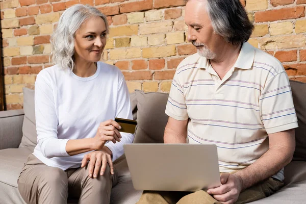 Mujer feliz y madura con tarjeta de crédito cerca de la computadora portátil y marido de mediana edad - foto de stock