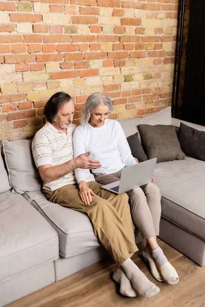 Hombre de mediana edad señalando con la mano a la computadora portátil cerca de la esposa - foto de stock