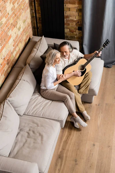 Blick von oben auf einen bärtigen und reifen Mann, der neben seiner fröhlichen Frau Akustikgitarre spielt — Stockfoto