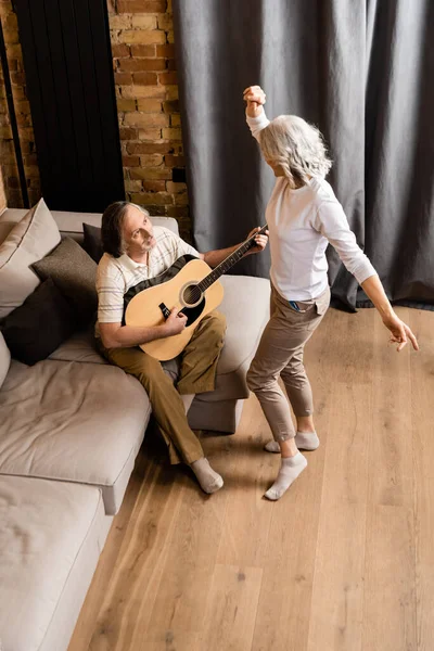 Barbudo y maduro hombre tocando la guitarra acústica y mirando alegre esposa bailando en sala de estar - foto de stock