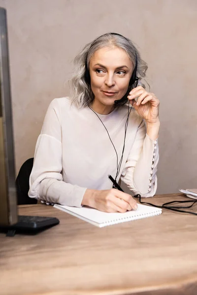 Selective focus of mature operator touching headset and writing in notebook near computer monitor at home — Stock Photo