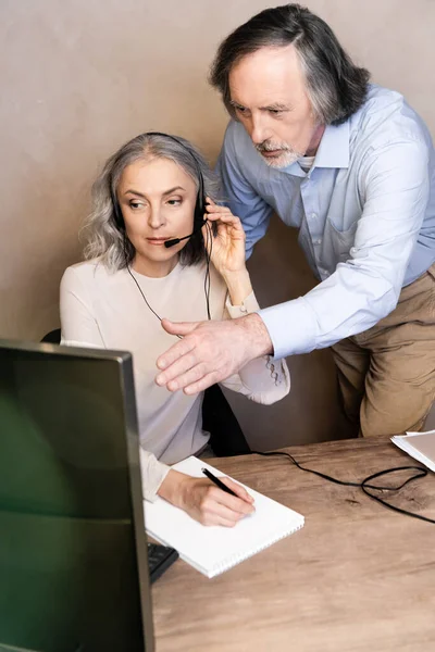 Enfoque selectivo del operador maduro tocando auriculares cerca de marido señalando con la mano en el monitor de la computadora - foto de stock