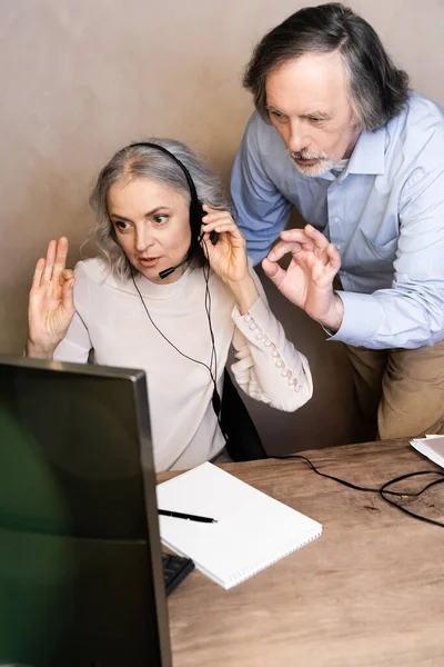 Foyer sélectif de l'opérateur mature dans le casque parlant près du mari montrant ok signe — Photo de stock