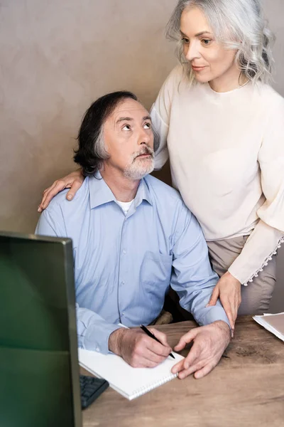 Foco seletivo de homem maduro escrevendo em notebook e olhando para a esposa perto do monitor de computador — Fotografia de Stock