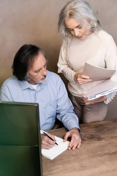 Selektiver Fokus des reifen Mannes, der in Notizbuch in der Nähe seiner Frau schreibt, die Ordner hält — Stockfoto