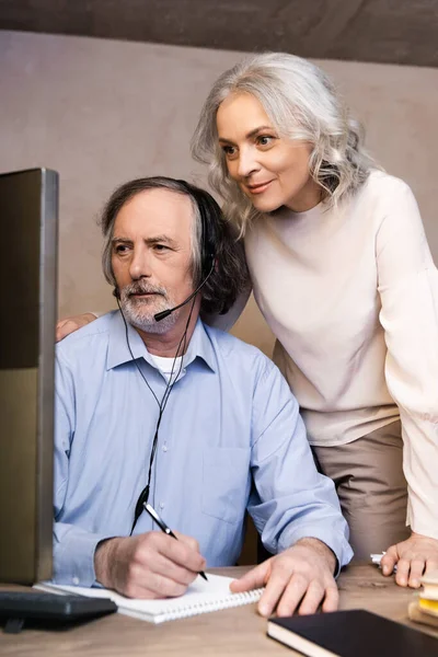 Selective focus of middle aged operator in headset looking at computer monitor near wife — Stock Photo