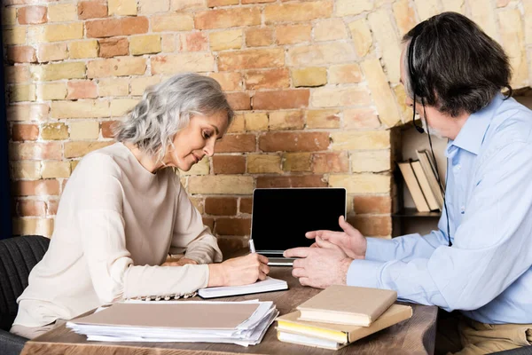 Mature woman writing near laptop with blank screen and husband in headset — Stock Photo