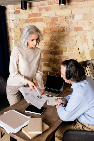 Femme mature tenant des graphiques et des graphiques près de mari dans un casque et un ordinateur portable avec écran blanc — Photo de stock