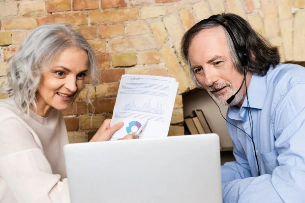 Mulher madura feliz segurando gráficos e gráficos perto do marido no fone de ouvido e olhando para o laptop — Fotografia de Stock