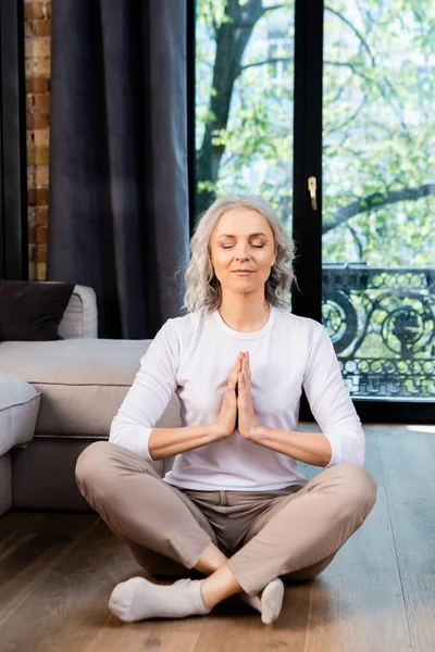 Relaxed mature woman with closed eyes and praying hands sitting in lotus pose — Stock Photo