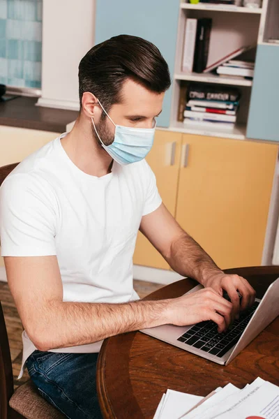 Людина в медичній масці використовує ноутбук біля паперів на столі вдома — стокове фото