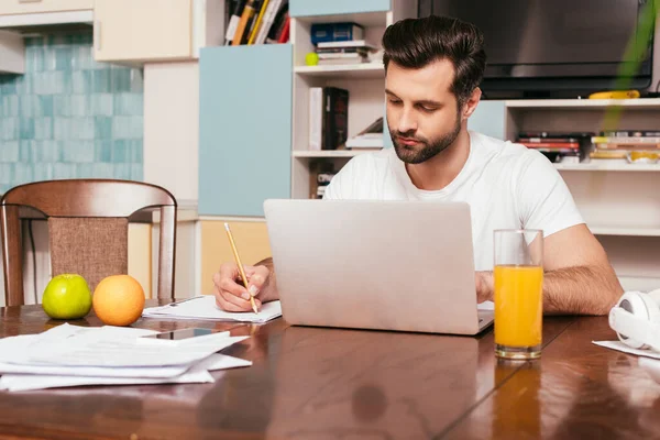 Foco seletivo de freelancer bonito trabalhando com papéis e laptop perto de suco de laranja e frutas na mesa — Fotografia de Stock