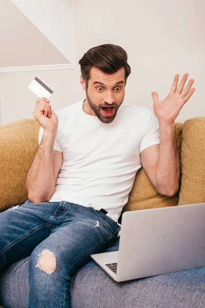 Hombre sorprendido mirando a la computadora portátil mientras sostiene la tarjeta de crédito en el sofá - foto de stock