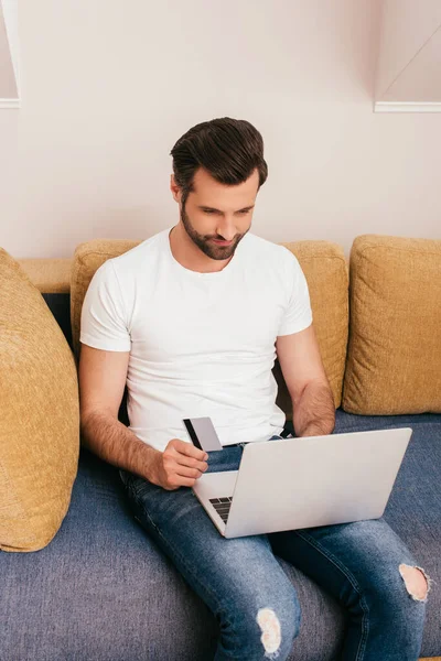 Bonito homem usando laptop e cartão de crédito no sofá — Fotografia de Stock