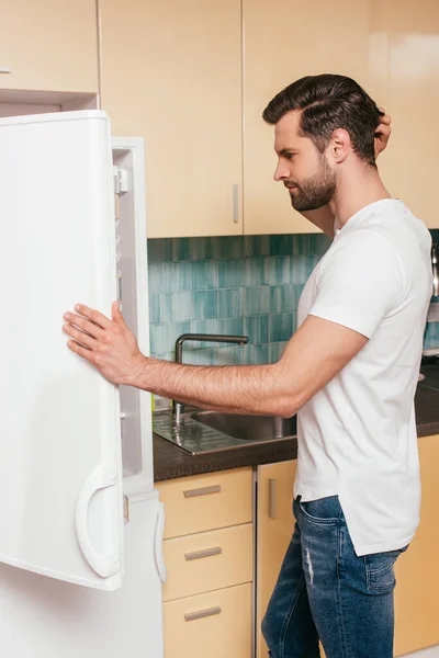 Side view of pensive man looking at open fridge in kitchen — Stock Photo