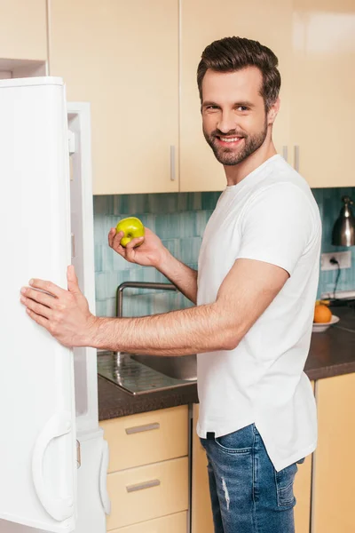 Side view of handsome man smiling at camera while holding apple near open fridge — Stock Photo
