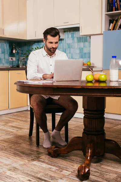 Красивый человек в рубашке и трусиках с помощью ноутбука рядом фрукты и молоко на кухне — стоковое фото