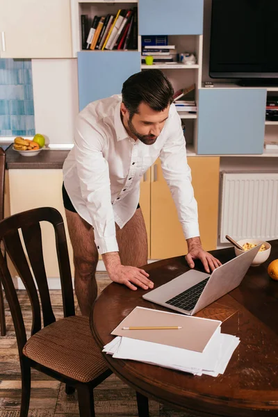 Bonito homem de camisa e calcinha usando laptop perto de cereais na mesa na cozinha — Fotografia de Stock