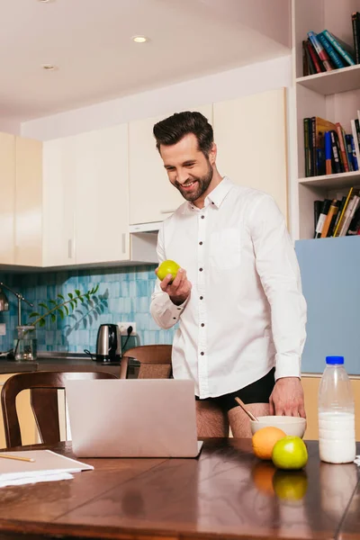 Selective focus of handsome man in panties and shirt smiling while holding apple near laptop on kitchen table — Stock Photo