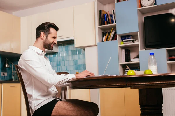 Vista laterale di uomo sorridente in camicia e mutandine utilizzando computer portatile vicino a colazione sul tavolo in cucina — Foto stock