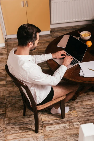 Vista aérea del hombre en bragas y camisa usando el ordenador portátil cerca de papeles y cereales en la mesa - foto de stock