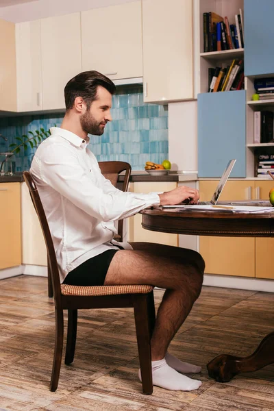 Side view of handsome freelancer in panties and shirt using laptop on kitchen table — Stock Photo