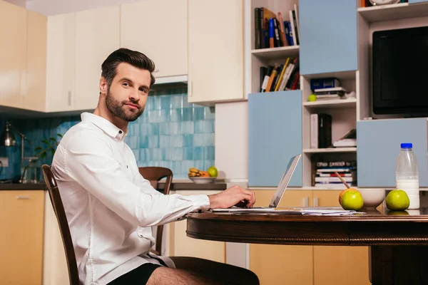 Vista lateral de belo freelancer em camisa e calcinha olhando para a câmera perto de laptop e café da manhã na mesa da cozinha — Fotografia de Stock