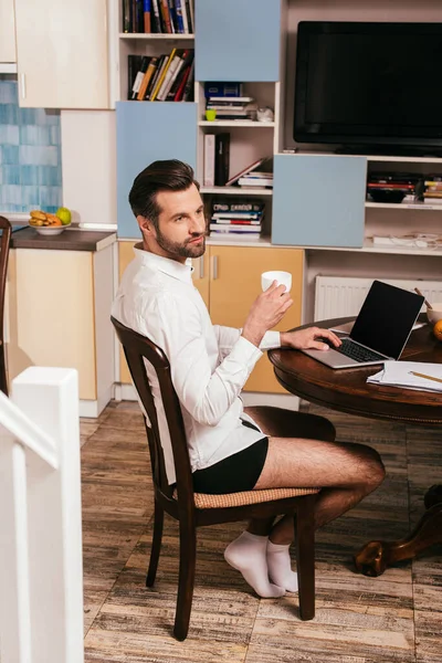 Focus sélectif de bel homme en chemise et culotte à l'aide d'un ordinateur portable et tenant la tasse dans le salon — Photo de stock