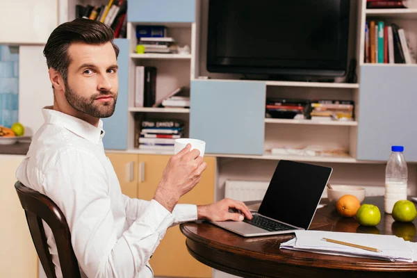 Vista lateral del hombre guapo sosteniendo taza de café cerca de la computadora portátil y papeles en la mesa - foto de stock