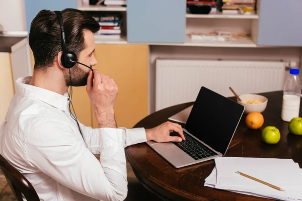 Vista laterale dell'uomo in cuffia che lavora su laptop vicino alla colazione sul tavolo — Foto stock