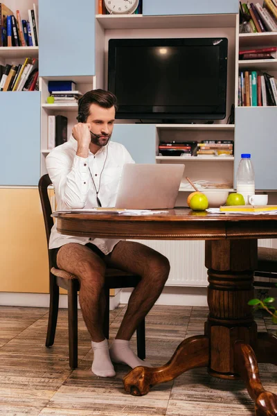 Freelancer em camisa e calcinha usando fone de ouvido e laptop perto do café da manhã na mesa da cozinha — Fotografia de Stock