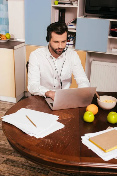 Красивий чоловік в гарнітурі використовує ноутбук біля сніданку на столі — стокове фото