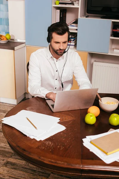 Handsome man in headset using laptop near breakfast on table — Stock Photo