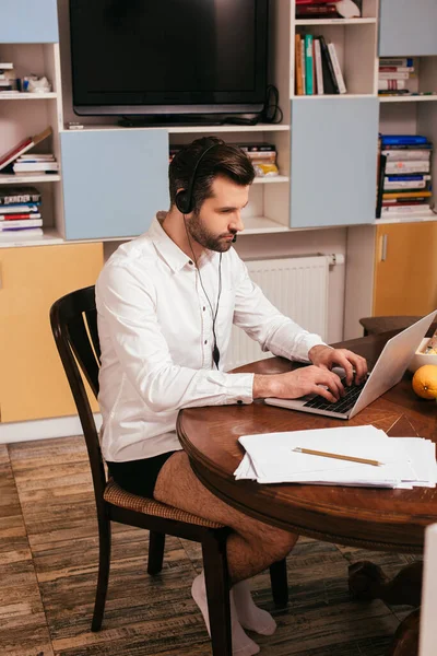 Enfoque selectivo del freelancer en camisa y bragas usando auriculares y portátil cerca de papeles en la mesa en casa - foto de stock