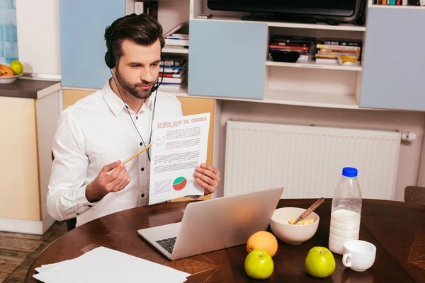 Freelancer en auriculares apuntando en papel con gráficos mientras tiene video chat en el portátil en casa - foto de stock