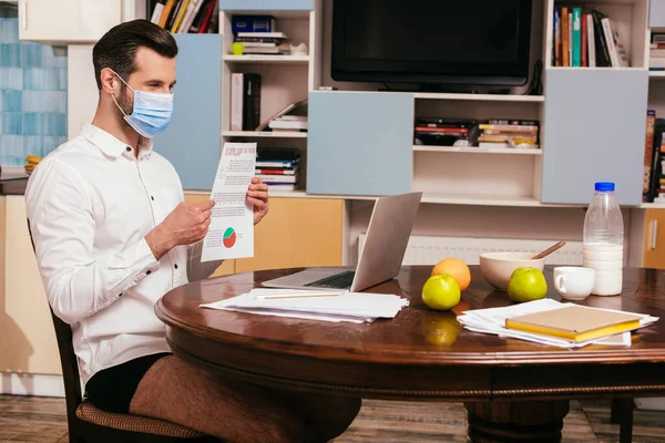 Freelancer en máscara médica, camisa y bragas que sostiene el documento mientras tiene video chat en el portátil cerca del desayuno en la mesa - foto de stock