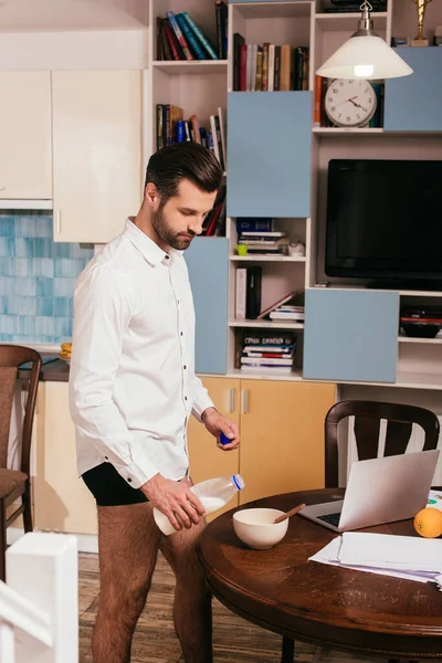 Vista lateral del hombre guapo en bragas y camisa vertiendo leche en un tazón cerca de la computadora portátil y papeles en la mesa - foto de stock