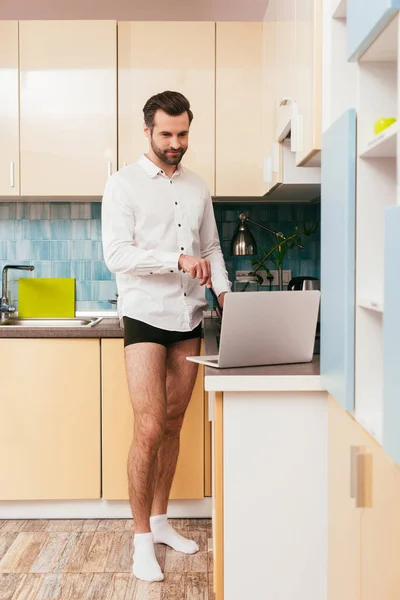 Bonito homem de camisa e calcinha olhando para laptop enquanto cozinha café da manhã na cozinha — Fotografia de Stock