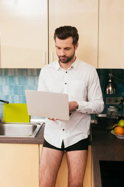 Handsome freelancer in shirt and panties using laptop in kitchen — Stock Photo