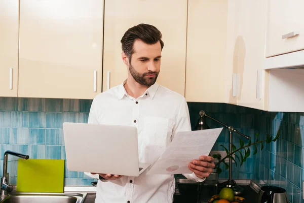 Hombre guapo mirando documento y sosteniendo el ordenador portátil en la cocina - foto de stock