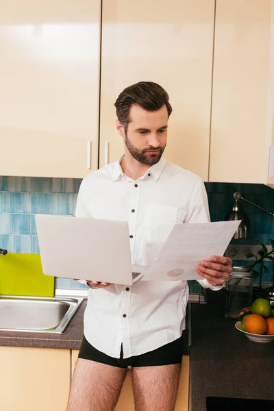 Bonito homem de camisa e calcinha trabalhando com documento enquanto segurando laptop na cozinha — Fotografia de Stock