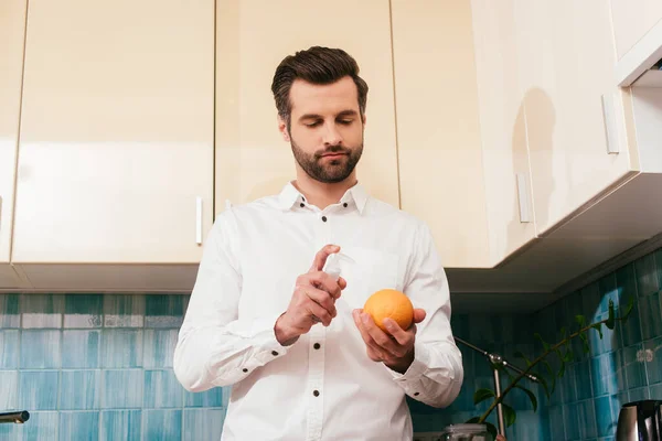 Vista de ángulo bajo del hombre guapo que sostiene el desinfectante naranja y de mano en la cocina - foto de stock