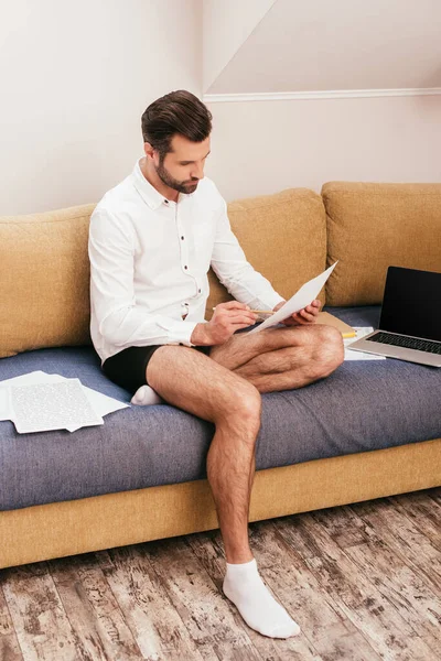 Handsome teleworker in panties and shirt working with papers near laptop on couch — Stock Photo