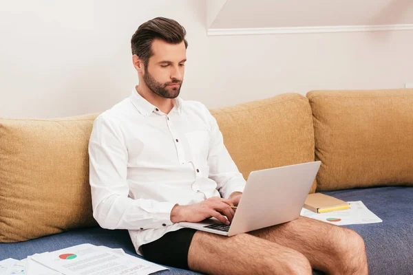 Teleworker in shirt and panties using laptop near papers on sofa in living room — Stock Photo