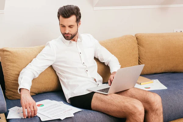 Freelancer en bragas y camisa usando laptop y trabajando con papeles en sofá en casa - foto de stock