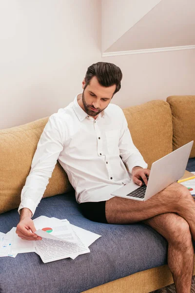 Handsome teleworker in panties and shirt working with documents and laptop on couch — Stock Photo