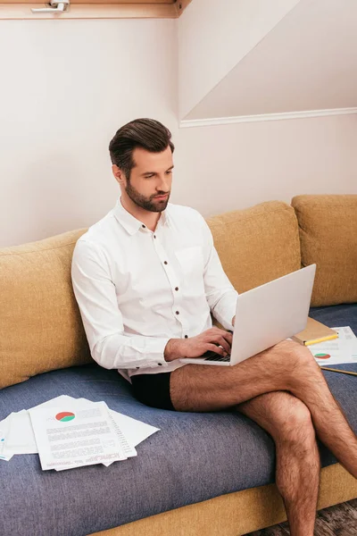 Bonito homem de camisa e calcinha usando laptop perto de papéis e livro no sofá — Fotografia de Stock