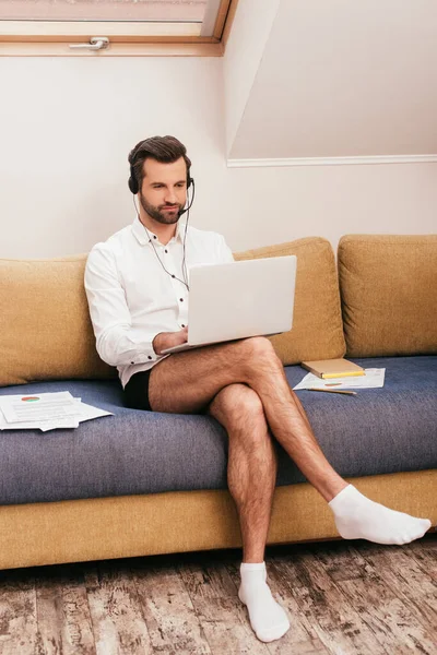 Handsome man in shirt and panties using laptop and headset on couch at home — Stock Photo