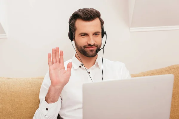 Selective focus of smiling freelancer in headset having video call on laptop on couch — Stock Photo